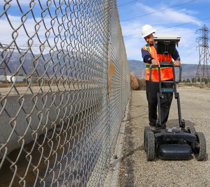 Worker using ground-penetrating radar near a fence in a construction area.
