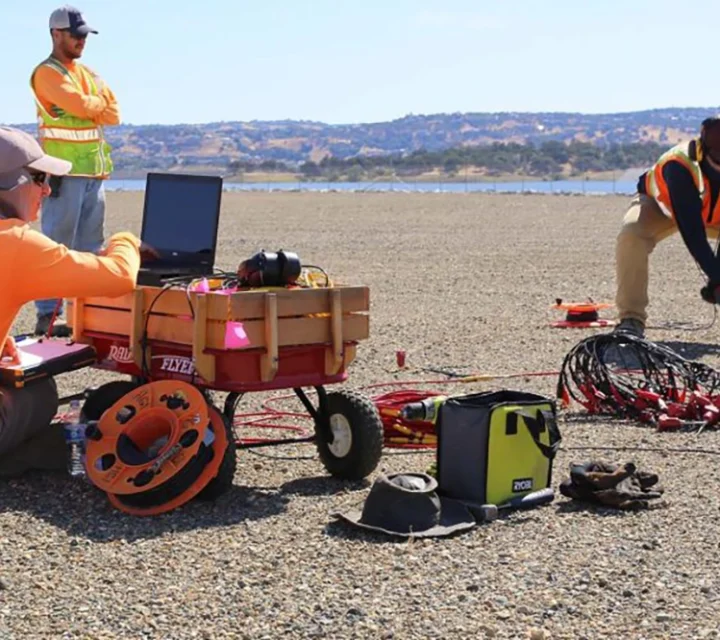 Survey crew using seismic equipment on rocky terrain near a lake.