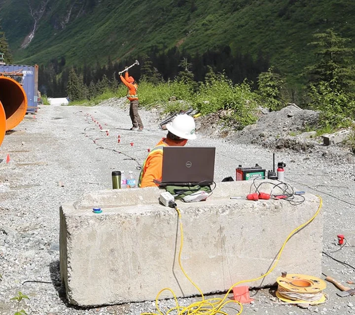 Surveyors working with equipment on a gravel road near large pipes and a forested hillside.