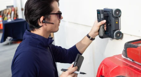 Man using handheld scanner on wall, holding notebook and pen, wearing headset and sunglasses.