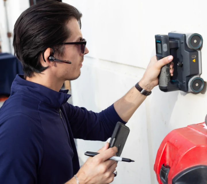 Man using handheld scanner on wall, holding notebook and pen, wearing headset and sunglasses.